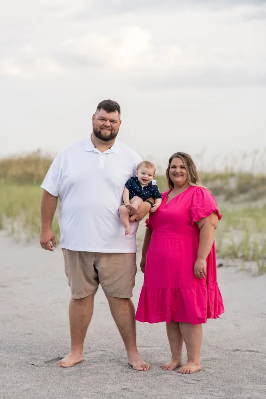 Josh and Brook stand together on the beach holding their young son, with dune grass behind them.