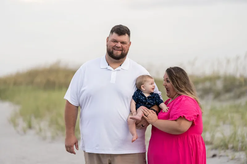 Josh holds their toddler while Brook stands beside them in a bright pink dress at the shoreline.