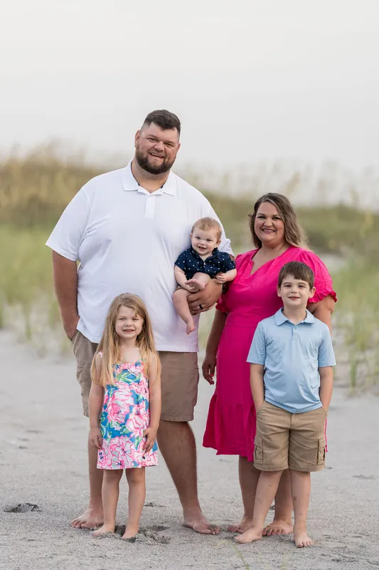 Brook and Josh stand with their three children on the sand at Vilano Beach, all smiling at the camera under an overcast sky.