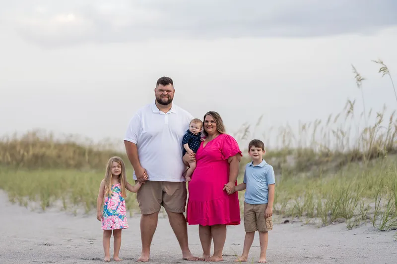 The family of five poses together among beach grasses, with their toddler held in Josh's arms and two older children standing