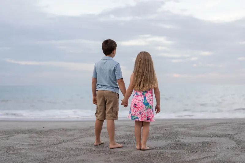Two children hold hands and wade toward the ocean at the shoreline, their backs to the camera against a cloudy horizon.