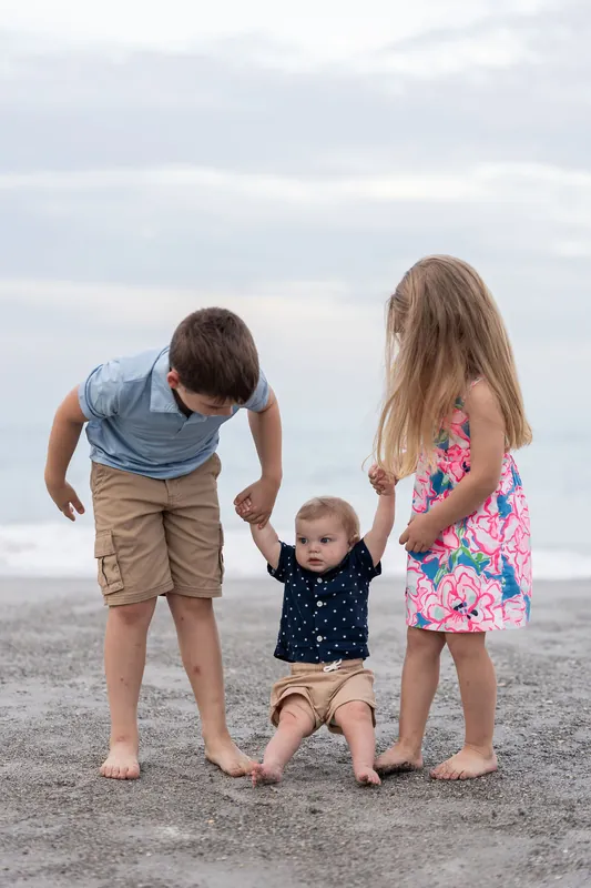 Three children stand together on the sand—two older kids flanking their toddler sibling in the middle—all looking down