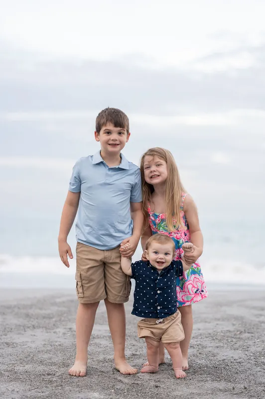 Three children stand barefoot on the sand at Vilano Beach, with a boy in light blue, a girl in a floral dress, and a toddler