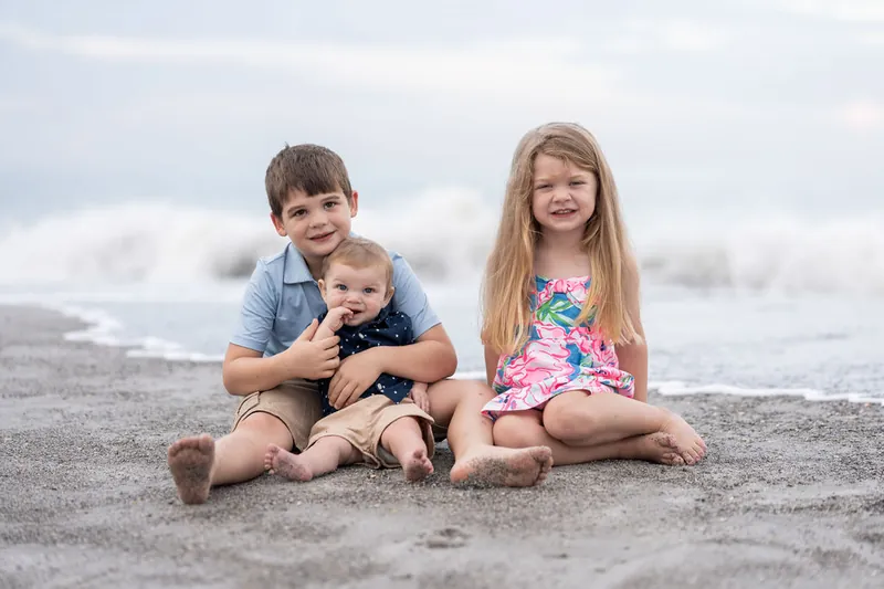 Two older children sit together on the beach while cradling their youngest sibling between them, ocean waves visible behind