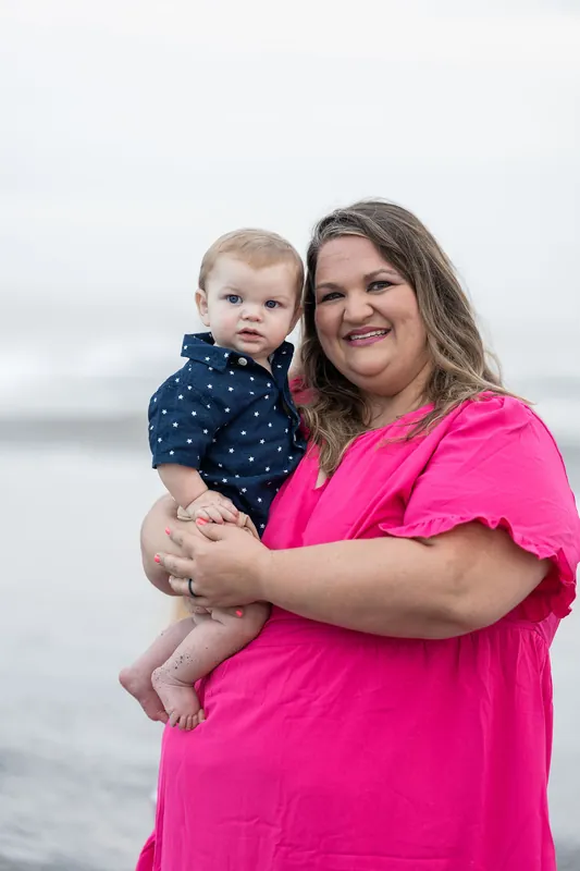 Brook holds their toddler wearing a navy polka-dot outfit while smiling at the camera in a bright pink shirt.