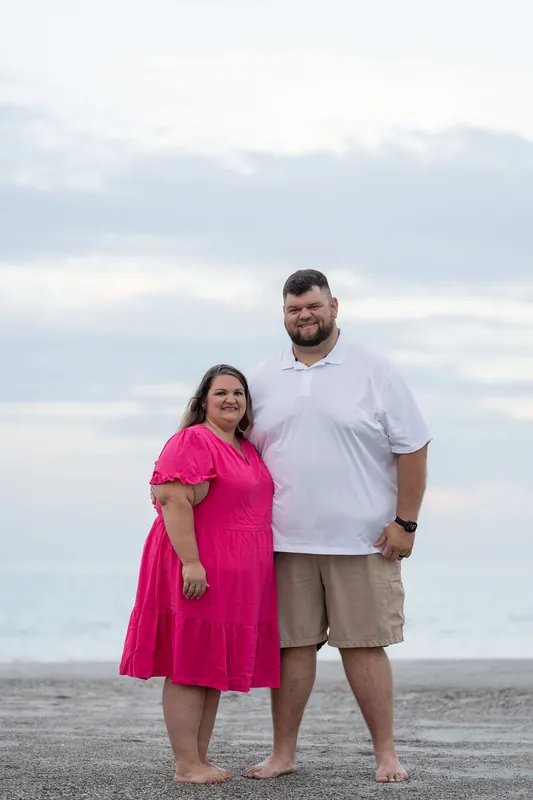 Josh and Brook stand together on the sand at Vilano Beach under an overcast sky, Brook in hot pink and Josh in white and tan.