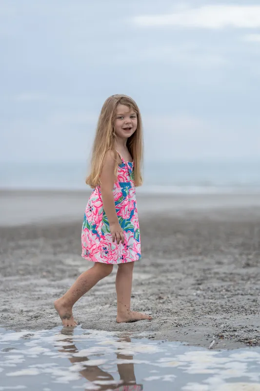 A girl in a colorful floral dress walks through shallow water at the shoreline, looking toward the camera.