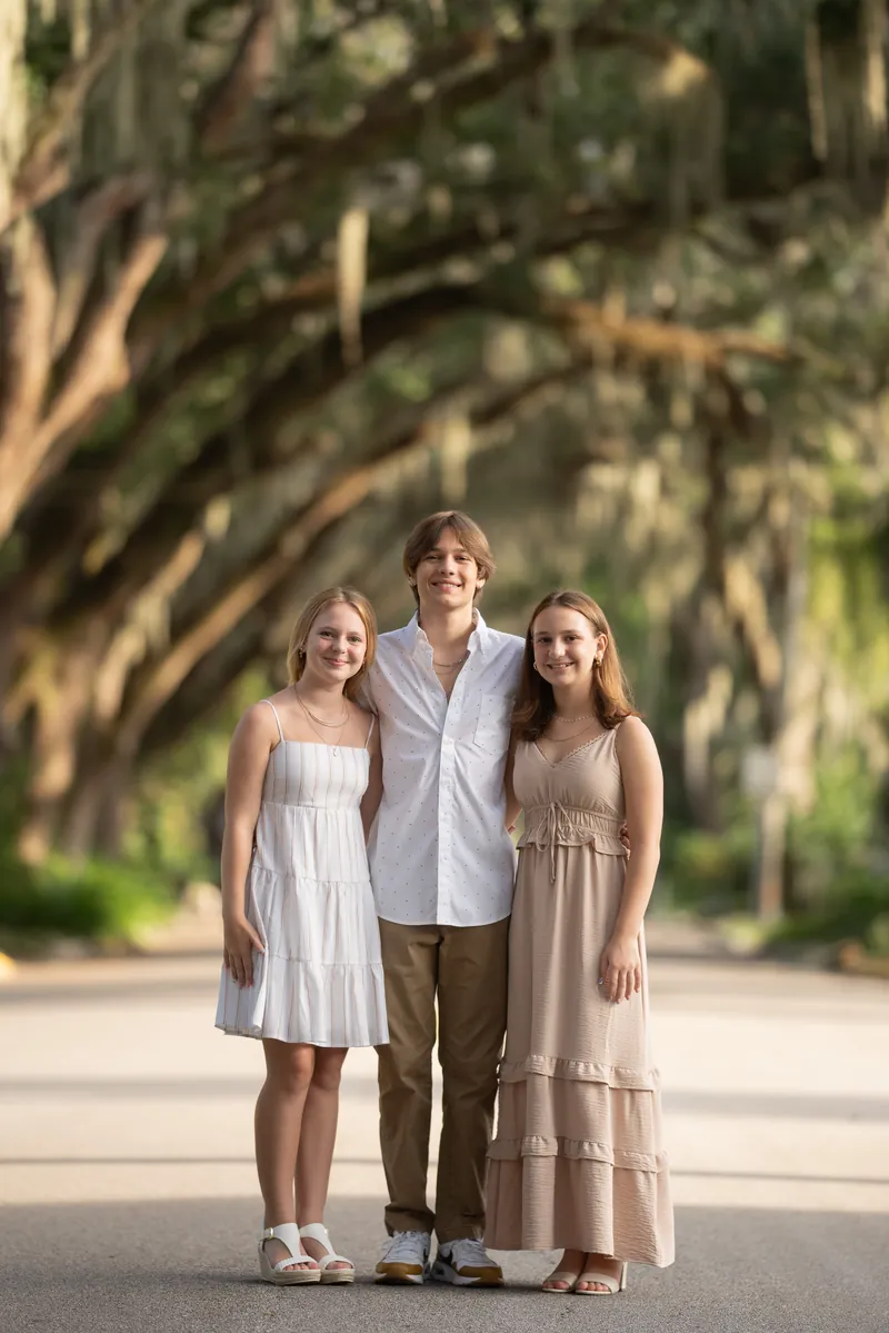 Three siblings standing together under oak canopy - brother in center with sisters on each side