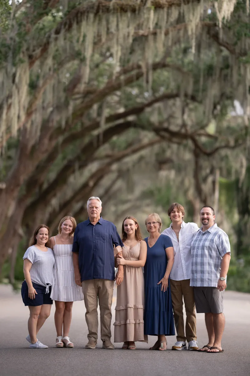 Eight family members stand together on a tree-lined street beneath sprawling oak branches draped with Spanish moss.