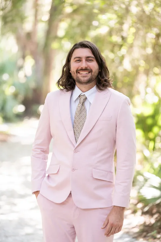 Cameron stands alone in a pink suit and gray tie, smiling at Fort Mose Boardwalk beneath dappled tree shade.