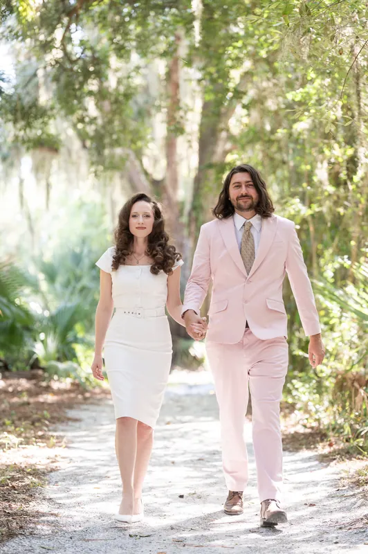 Cameron and Chris walk hand-in-hand along the boardwalk, sunlight filtering through the dense tree cover overhead.