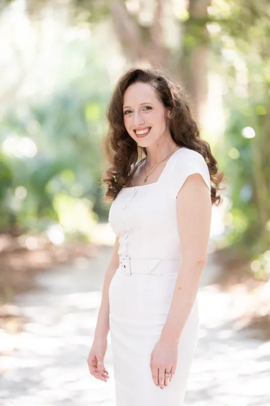 Person in white cap-sleeve dress smiling at the camera beneath tree-lined boardwalk at Fort Mose.