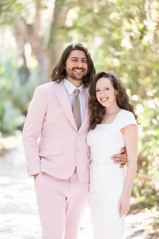Cameron and Chris stand close together on the boardwalk, Cameron in pink suit and Chris in white dress.