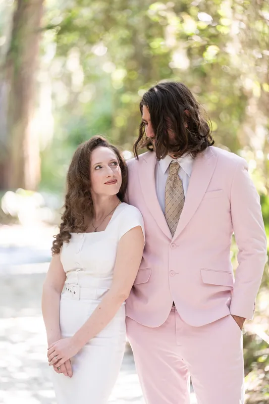 A person in a pink suit holds hands with their partner in a white dress under the oak trees.