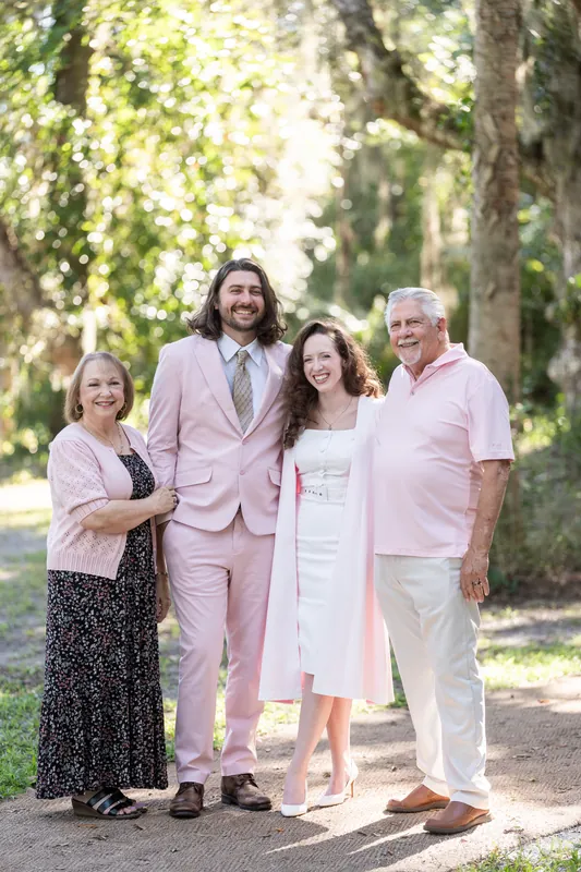Four people stand together on a shaded boardwalk lined with tall trees at Fort Mose Boardwalk, St. Augustine, wearing pastels and light neutrals.