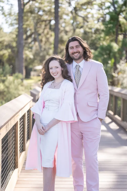 Two people in pastel suits stand arm-in-arm on a wooden boardwalk at Fort Mose, one in pink and one in white.