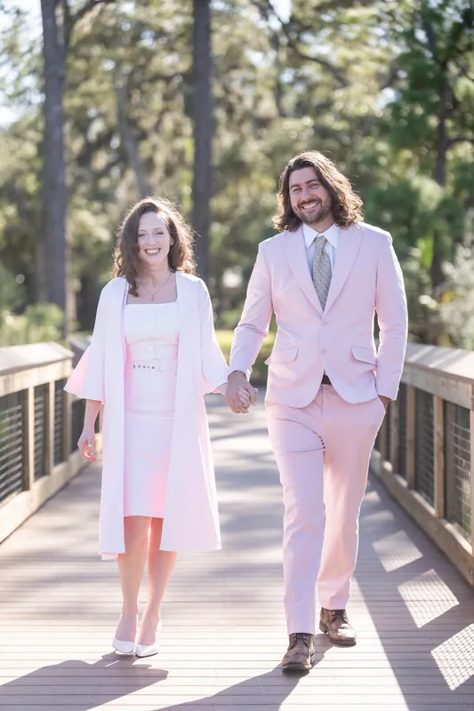 The couple walks hand-in-hand down the sun-dappled boardwalk at Fort Mose, Cameron in pink and Chris in white, with trees framing the path ahead.