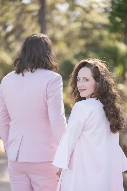 Two people in pastel wedding attire stand back-to-back on the shaded boardwalk, one in a pink suit and one in a white dress.