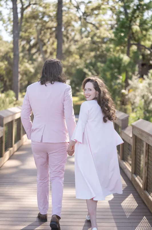 Cameron and Chris walk away from the camera down the wooden boardwalk, their silhouettes framed by tall trees and filtered sunlight at Fort Mose.