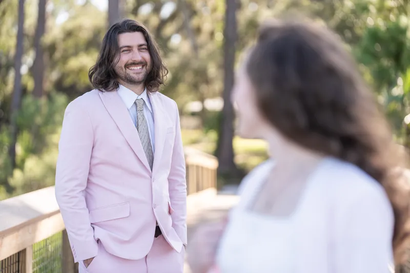 A person in a pink suit smiles across the boardwalk at someone in a white dress, with trees and dappled light behind them at Fort Mose.