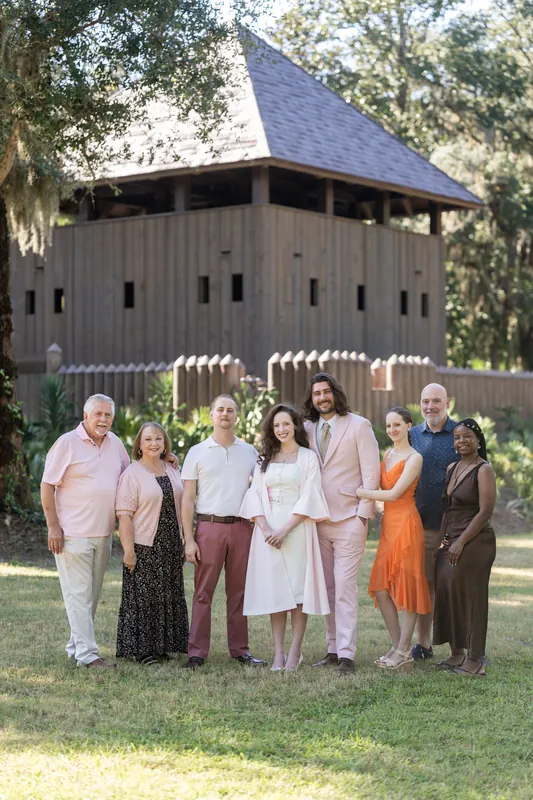 Eight people in mixed casual and formal wear stand on grass before Fort Mose's wooden structure, framed by moss-draped oaks.
