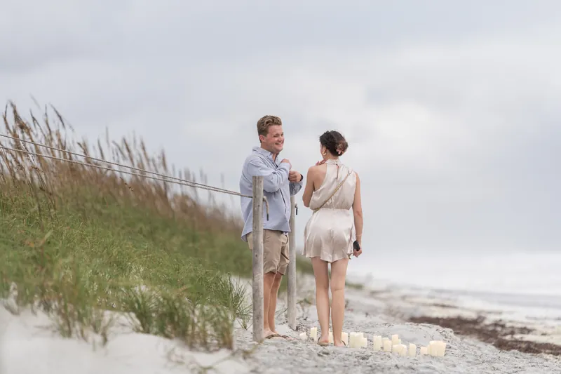 Two people stand close together on sand beside dune grass at Mussallem Beachfront Park, ocean and grey sky behind them.