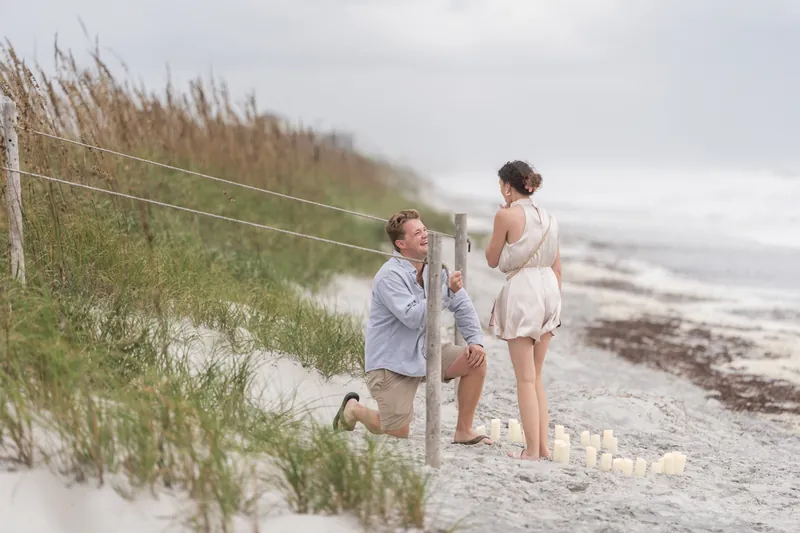 A person kneels on the sand while another stands nearby as the proposal unfolds at Mussallem Beachfront Park, St. Augustine.