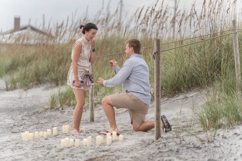 One person kneels on the sand holding a ring box while the other stands nearby during the proposal moment.