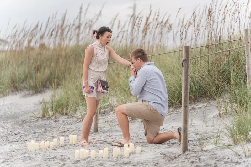 Two people embrace on the sand after the proposal, with beach dunes and fence visible behind them.