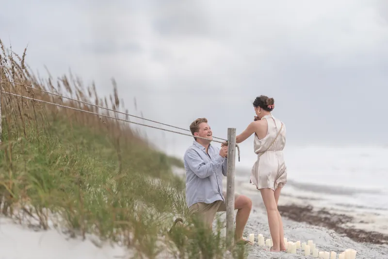 A newly engaged couple celebrates together on the beach with dunes and ocean visible in background.