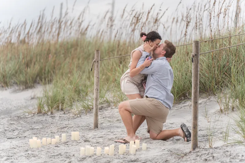 Carter kneels on the sand at Mussallem Beachfront Park beside candles arranged in a heart, holding Emily's hand as she stands in a white dress.