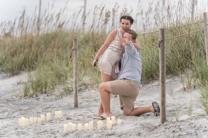 Emily leans toward Carter as he kneels on the sand, both smiling, with dune grass and wooden fence posts framing the moment at the beach.