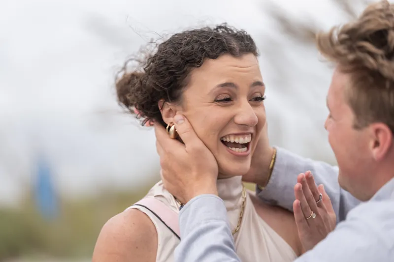 A close-up captures Emily's radiant smile and wide eyes as she looks toward Carter, her hand raised near her ear at the beachfront.