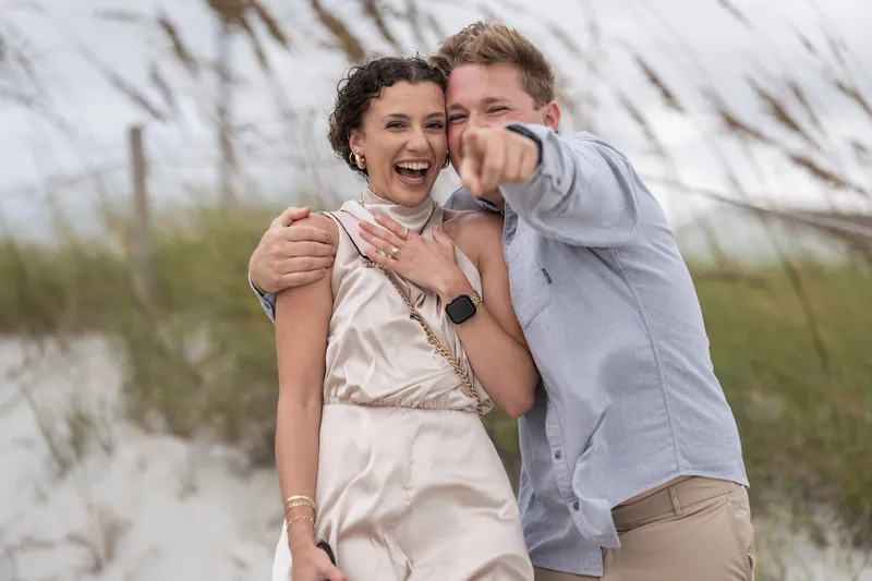 Carter and Emily embrace on the sand with the dune landscape behind them, both beaming with Emily holding her hand near her heart.