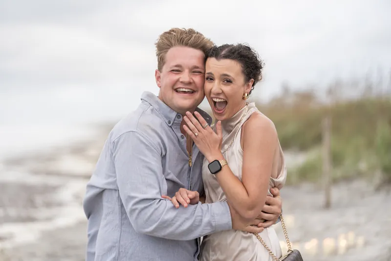 The couple stands together on the beach laughing, Emily displaying her ring hand while Carter wraps his arm around her at Mussallem Beachfront Park.