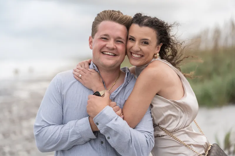 Two people hug and smile broadly on the sand with dune grasses and water visible behind them.