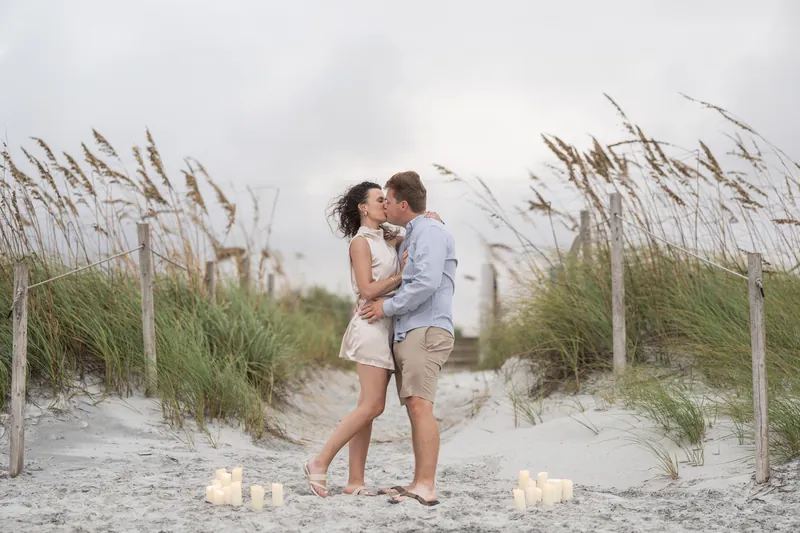 A couple kisses surrounded by beach dunes and sea oats, with candles glowing nearby in the sand.