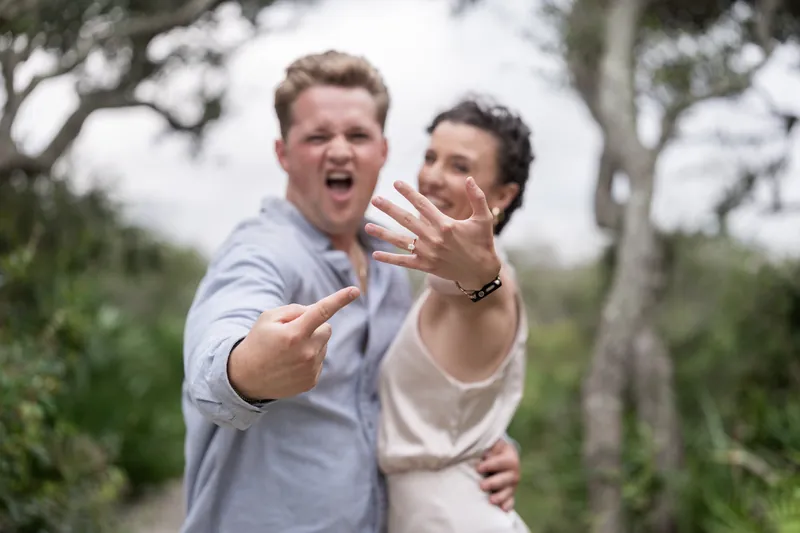 Two people display their hands showing off a new engagement ring while laughing beneath oak trees.