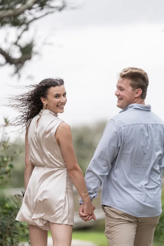 Two people hold hands and walk together along a grassy area framed by large oak trees overhead.