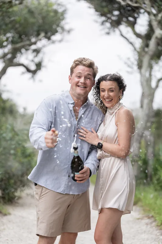Carter and Emily celebrate with champagne flutes among live oak trees at Mussallem Beachfront Park in St. Augustine.