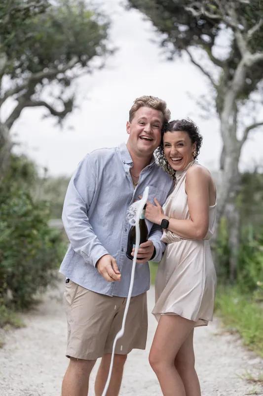 The couple clinks glasses together on a tree-lined path, both smiling broadly at the beachfront park.