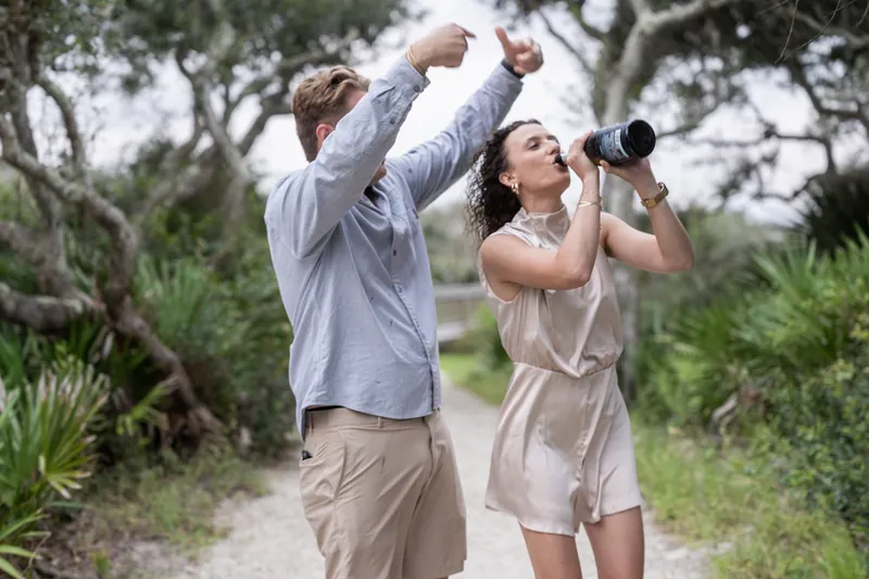 Each holds binoculars toward the sky along a sandy trail framed by gnarled oaks and palmetto vegetation.