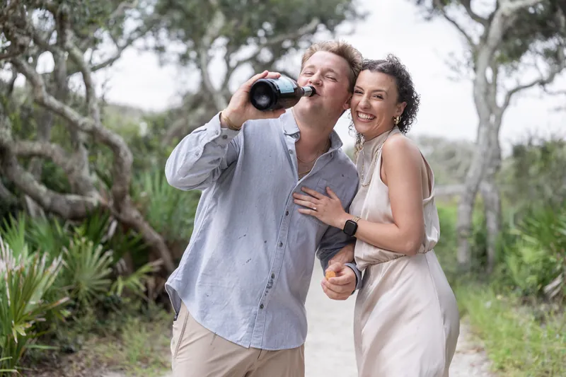 One person raises binoculars while the other watches, both holding champagne bottles amid the coastal landscape.