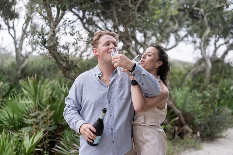 Someone takes a sip from a champagne flute as their partner looks on with a smile among the park's trees.