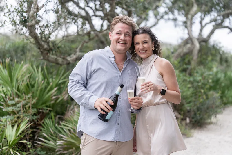 The pair stands close together holding champagne bottles on a sandy path bordered by live oaks at the beachfront venue.