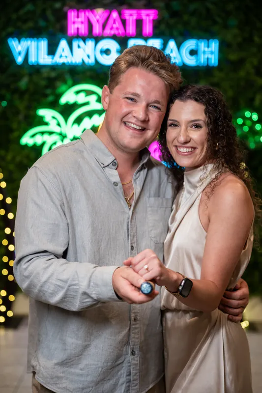 Carter and Emily stand smiling beneath neon signage for Hyatt Vilano Beach, each holding the compass proposal token.