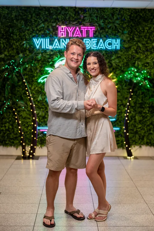 Carter and Emily pose together on an outdoor patio lit by greenery and warm string lights at the beachfront venue.