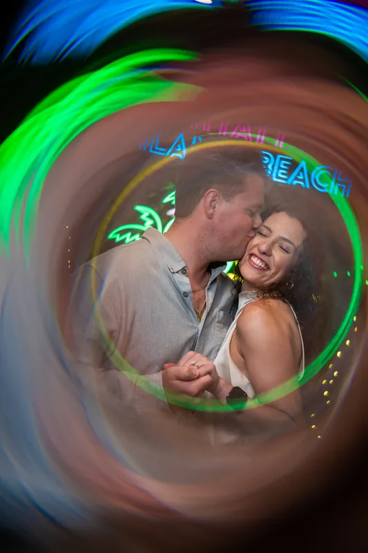 The couple embraces within a swirl of glowing neon light patterns reflecting off the wet ground around them.