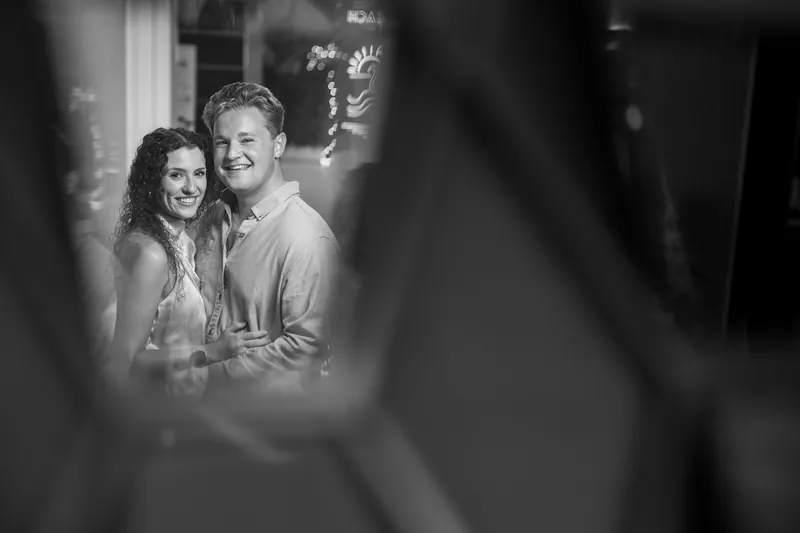 A black and white portrait shows two people gazing at each other through sheer fabric in soft indoor lighting.