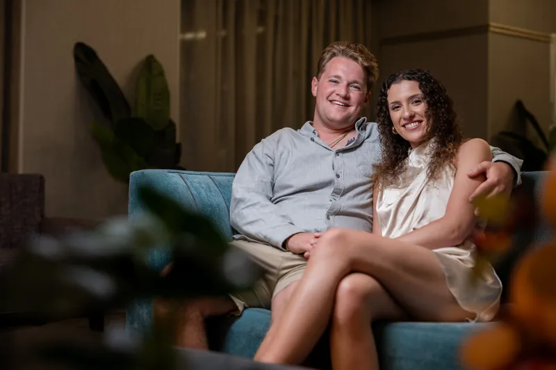 Carter and Emily sit close together on a blue couch surrounded by potted plants, smiling warmly at each other indoors.